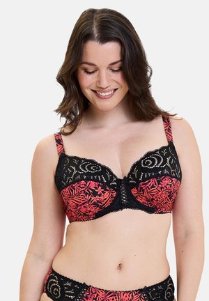 Donna sorridente che indossa un reggiseno in pizzo con motivo floreale rosso e nero e biancheria intima coordinata, con capelli castani ondulati e che guarda verso il basso.