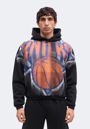 Jeune homme portant un sweat à capuche noir avec un grand ballon de basket orange et le mot "KNICKS" en lettres majuscules sur le devant.