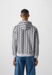 Felpa con cappuccio grigia con motivo geometrico nero. Presenta una superficie morbida e un orlo a coste. Abbinata a pantaloni cargo blu.