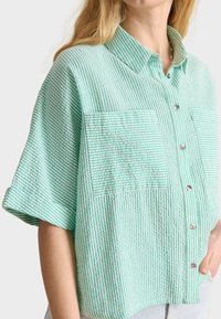 Short-sleeve, collared shirt in light green with white vertical stripes, featuring two front pockets with textured fabric and silver buttons.