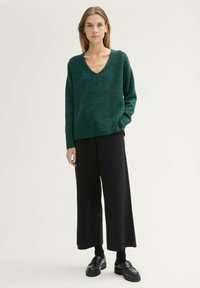 Pulover tricotat verde cu guler în formă de v, cu mâneci lungi, asortat cu pantaloni negri largi și mocasini negri masivi, pe un fundal simplu.