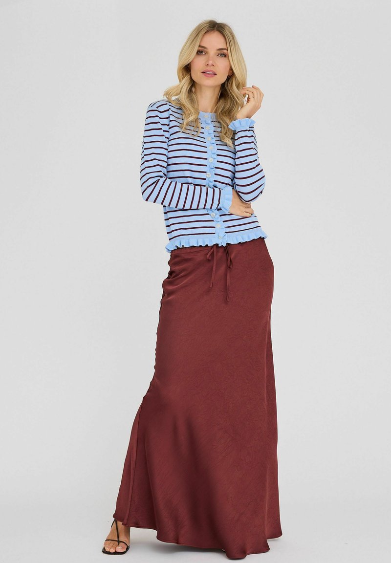 Femme aux cheveux blonds portant un cardigan à rayures bleues et une longue jupe marron, debout devant un fond uni gris clair.