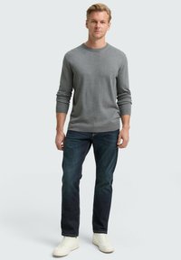 Man staat in een grijze lange mouw sweater, donkere spijkerbroek en witte sneakers tegen een effen lichte achtergrond.