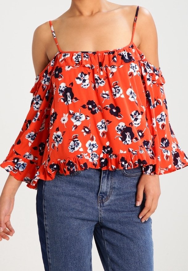 Femme portant un chemisier floral orange à épaules dénudées avec des volants, associé à un jean taille haute bleu.