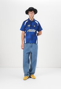 Blue polo shirt with white accents, denim jeans, and yellow sneakers. Model wearing a black bucket hat. Features logo and stylized design.