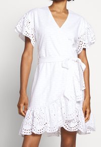 Abito avvolgente bianco con maniche corte a volant, motivi a occhio di pernice, scollatura a V, vita legata e orlo a balze.