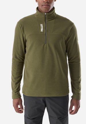 Man wearing an olive green long-sleeve half-zip fleece top and dark gray pants, standing against a white background.