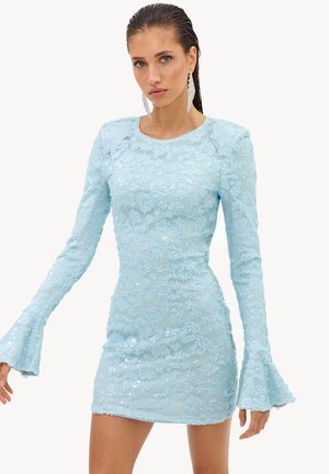 Femme portant une mini-robe moulante bleu clair à sequins, à manches longues, avec poignets évasés et grandes boucles d’oreilles pendantes, debout devant un fond uni.