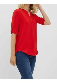 Rød kortærmet bluse med V-hals, tekstureret stof, opsmøgede ærmer og en let buet kant, parret med mørkeblå jeans.