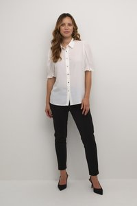 Blusa bianca con maniche a sbuffo, chiusura con bottoni e rifiniture in pizzo, abbinata a pantaloni neri aderenti e scarpe con tacco nere.