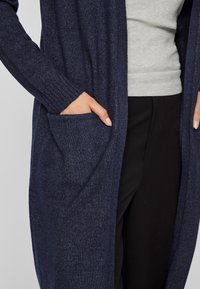 Ναυτικό πλεχτό cardigan με ριγωτούς καρπούς, ανοιχτό σχέδιο μπροστά και δύο πλαϊνές τσέπες. Φορεμένο πάνω από γκρι τοπ και μαύρα παντελόνια. Μαλακή υφή.