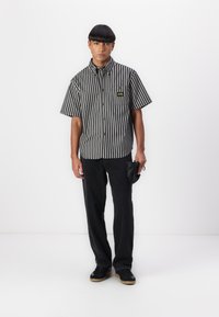 Short-sleeve black and white striped button-up shirt with a chest pocket; paired with black pants and black sneakers.