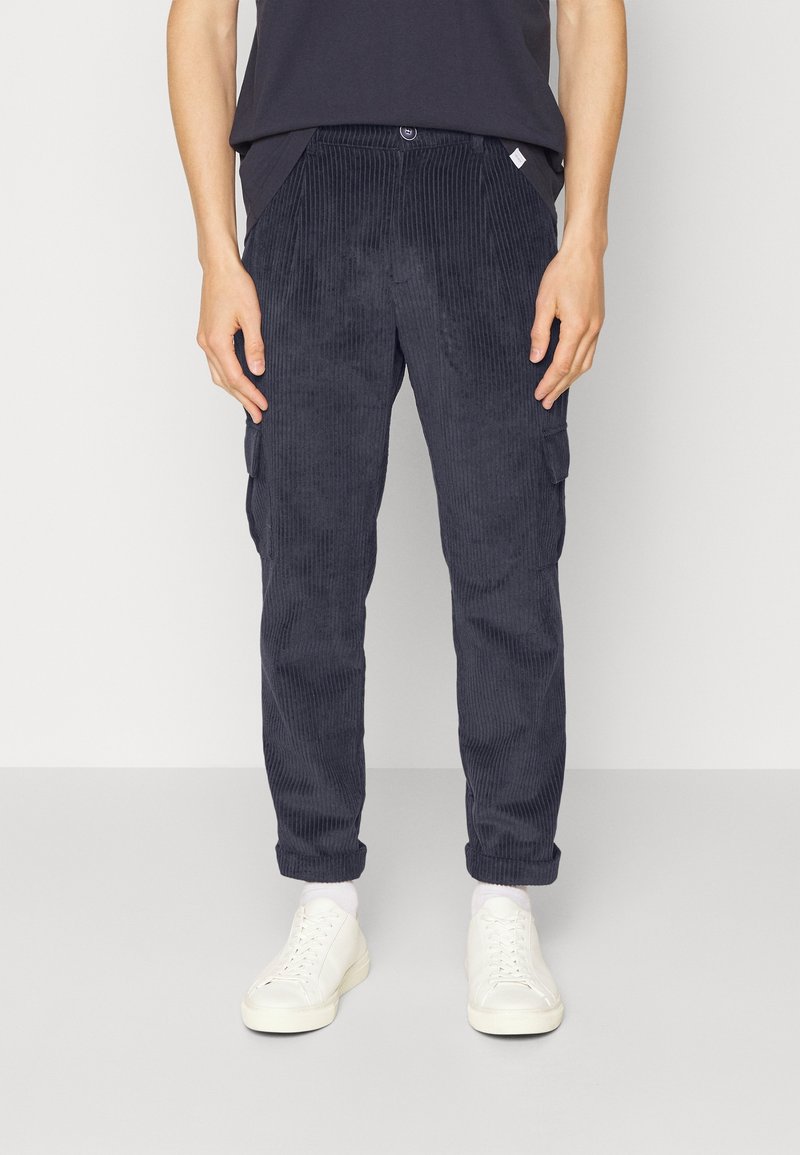 Pantalon cargo en velours côtelé bleu marine avec poches latérales, coupe fuselée, tissu texturé et fermeture à boutons, associé à des baskets blanches.
