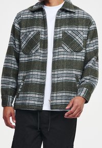 Giacca zip con motivo a quadri verde e bianco, colletto, due tasche sul petto e una texture morbida. Il design presenta motivi orizzontali e verticali.