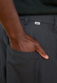 Dark gray pants with a textured finish, featuring a side pocket and a small white label reading "OBEY" near the waistband.