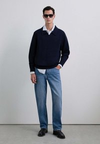 Suéter azul marino con un gran cuello, superpuesto sobre una camisa blanca con rayas. Jeanson de un tono azul claro y zapatos negros completan el conjunto.