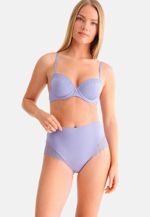 Vrouw draagt lavendelkleurige slip met hoge taille en bijpassende beugelbeha met kanten rand, staand tegen een effen lichte achtergrond.
