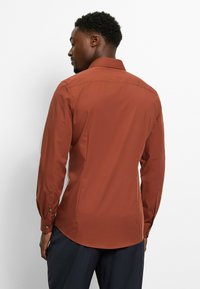 Rust-colored long-sleeve shirt with a fitted design, a classic collar, and buttoned cuffs. Smooth fabric texture, curved hemline at the back.
