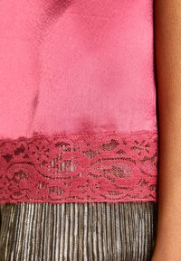 Tejido de satén rosa con un borde de encaje floral sobre una falda metálica plisada, junto a un brazo de tono de piel marrón.