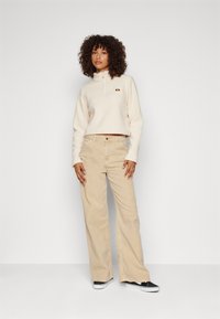 Kremainen lyhyt fleece-pullovaraios, jossa on puoliveto ja logo, yhdistettynä vaaleanbeigeihin leveälahkeisiin vaaleanpunaisiin housuihin ja mustiin liukuviin kenkiin.