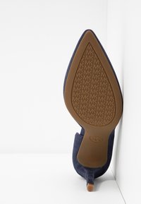 Chaussure à talon haut en daim bleu marine avec une pointe effilée, dotée d'une semelle en caoutchouc brun texturée embossée du logo MK.