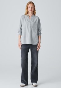 Camicia grigia a maniche lunghe con mezza abbottonatura e tasca sul petto, abbinata a jeans larghi scuri e sneaker bianche. Tessuto di trama liscia.