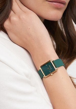 Grüner Silikonarmbanduhr mit einem quadratischen goldenen Gehäuse und dunkelgrünem Zifferblatt, das goldene Stundenmarkierungen und Zeiger aufweist.