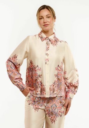 Žena u krem svilenoj bluzi i odgovarajućim hlačama s ružičastim i narančastim složenim cvjetnim paisley uzorcima, stoji s jednom rukom u džepu.