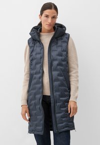 Navyblaues gepolstertes Vest mit Kapuze, mit Reißverschluss vorne und seitlichen Taschen, getragen über einem beigen Pullover und dunklen Jeans.
