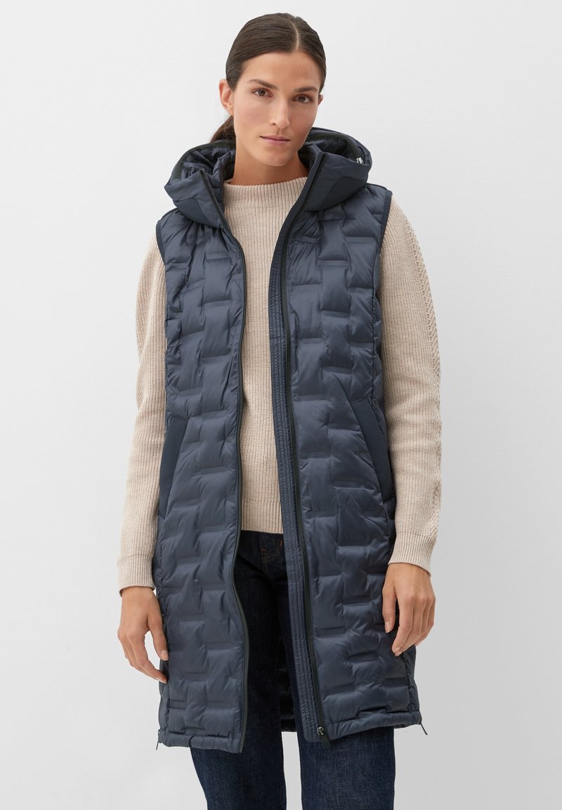 Navyblaues gepolstertes Vest mit Kapuze, mit Reißverschluss vorne und seitlichen Taschen, getragen über einem beigen Pullover und dunklen Jeans.