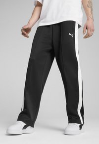 Sorte træningsbukser med hvide sidelinjer, lavet af glat materiale. Har et logo på låret og er parret med hvide sneakers.