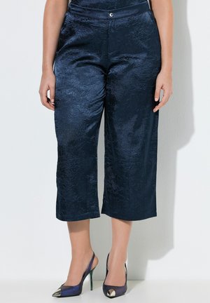 Frau trägt eng geschnittene, strukturierte marineblaue Hosen und metallisch spitz zulaufende Slingback-Pumps mit Stilettoabsätzen, steht vor einer weißen Wand.