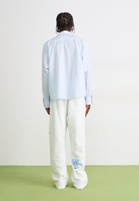 Person med fletninger iført en lys blå stribet langærmet skjorte og hvide sweatpants med blå grafik på underbenet står med ryggen til.