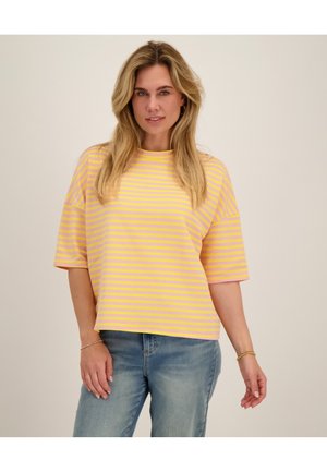 Vrouw met lang blond haar die een geel en roze gestreept T-shirt met korte mouwen en blauwe spijkerbroek draagt tegen een effen achtergrond.