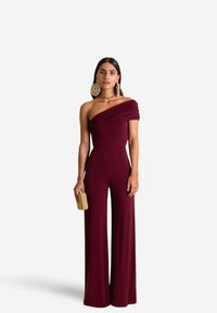 Jumpsuit bourgogne à une épaule avec des détails drapés, jambes larges et corsage ajusté. Le mannequin tient une pochette dorée texturée.