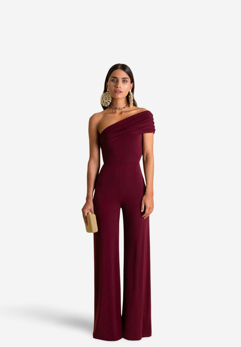 Jumpsuit bourgogne à une épaule avec des détails drapés, jambes larges et corsage ajusté. Le mannequin tient une pochette dorée texturée.