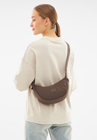 Borsa a spalla in tessuto marrone con forma curva, tracolla regolabile e patch con logo, indossata su una camicia chiara su uno sfondo neutro.