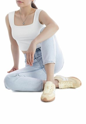 Zapatillas deportivas de color amarillo claro con acentos blancos, materiales de ante texturizado y malla, puntera redonda y suelas de goma color marrón claro en contraste, usadas con vaqueros de mezclilla claros.