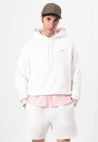 Sudadera blanca con bolsillo delantero y capucha con cordón, superpuesta sobre una camiseta de rayas rosas, combinada con pantalones cortos texturizados blancos y una gorra con estampado.
