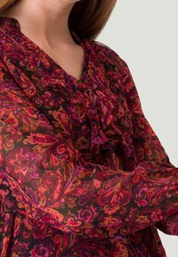Blouse transparente à manches longues en tissu sombre avec des motifs floraux et tourbillonnants complexes dans des nuances de rose, rouge et violet ; présente un col froncé.