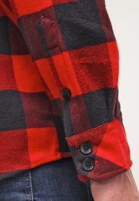 Manga de camisa de franela a cuadros rojos y negros con botones negros y un puño redondeado; la tela texturizada muestra patrones y colores contrastantes.