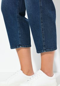 Tmavě modré zkrácené džínové kalhoty rovného střihu s obroubenými lemy, v kombinaci s bílými koženými teniskami.
