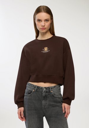 Felpa corta marrone con scollatura rotonda, maniche oversize e una grafica di un orso con testo sul petto. Indossata con jeans grigi a vita alta.