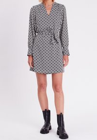 Robe noir et blanc avec un motif géométrique, manches longues, décolleté en V, et ceinture à nouer. Associée à des bottines noires.