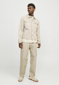 Beige denimjakke med to forlommer, parret med en lysefarvet lagdelt skjorte og beige cargobukser. Sneakers med cremefarvede og tan accenter.