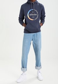Sudadera con capucha azul marino con cordón ajustable, que presenta un gráfico circular y el texto "Quiksilver". Combinada con pantalones vaqueros de puño azul claro y zapatillas blancas.
