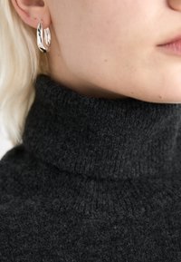 Brincos de argola em prata com um design torcido e texturizado, usados com um suéter de malha cinza de gola alta, mostrando um close-up dos materiais.