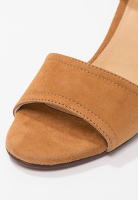 Gros plan d'une sandale ouverte en daim beige avec une large sangle avant et des coutures visibles sur fond blanc.