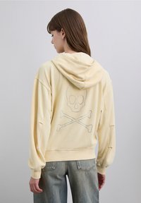 Gelber Hoodie aus weichem Stoff mit einem Strass-Schädel- und Knochenmotiv auf der Rückseite, mit lockerem Schnitt und Kapuze.