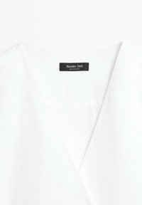 Vêtement blanc avec un col en V comportant une étiquette noire indiquant "Massimo Dutti Made in Morocco" cousue à l'intérieur du col.